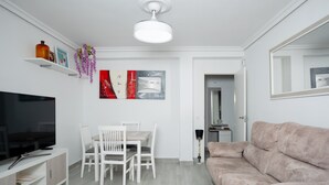 A modern living room with a flat-screen TV, white dining table, chairs, a beige sofa, and a ceiling fan.
