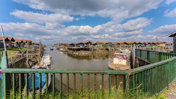 A riverside village with boats docked along the water, wooden houses with red roofs, and a green metal railing in the foreground. - Air-conditioned Zen Studio Near the Beaches, Quiet and Comfortable (Gujan-Mestras)