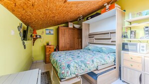 A compact bedroom with a loft-style ceiling, a bed with floral bedding, a wooden wardrobe, and a small kitchenette area. - Air-conditioned Zen Studio Near the Beaches, Quiet and Comfortable (Gujan-Mestras)