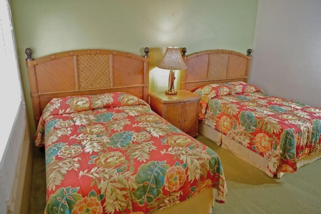 Een kamer met twee eenpersoonsbedden, elk voorzien van een bloemenprint deken, een houten hoofdbord en een nachtkastje met een lamp.