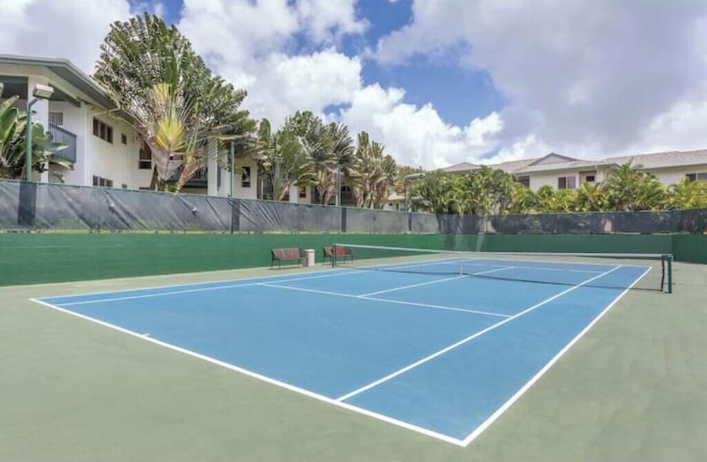 Ein Tennisplatz im Freien mit Netz, umgeben von einem grünen Zaun, neben einem Gebäude mit mehreren Fenstern und Balkonen.