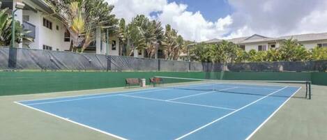 Ein Tennisplatz im Freien mit Netz, umgeben von einem grünen Zaun, neben einem Gebäude mit mehreren Fenstern und Balkonen.