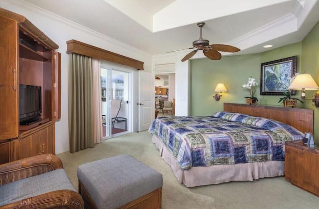 A hotel room with a large bed, a wooden wardrobe, a television, a ceiling fan, and a view of the adjacent room.