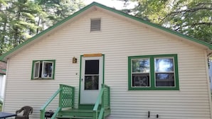 A small, single-story building with a green door and windows, a small porch with a bench, and a dirt ground area. - Hawthorne Cottages Chickadee Heights (Raymond)