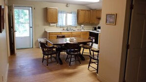 A kitchen with wooden cabinets, a dining table with chairs, a window with curtains, and a door leading to another room. - Hawthorne Cottages Chickadee Heights (Raymond)