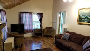 A living room with a brown sofa, a television, a wooden floor, and a window with curtains. - Hawthorne Cottages Chickadee Heights (Raymond)