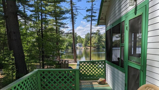 A wooden deck with green railings, a chair, and a view of a lake and a house in the background. - Hawthorne Cottages Sand Castle (Raymond)