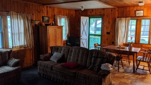 A cozy cabin interior with a wooden floor, a plaid sofa, a dining table, and a large window.