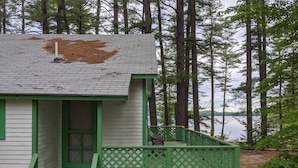 A small, single-story cabin with a green porch and a bench, surrounded by tall trees and a lake.
