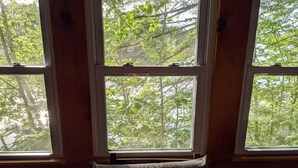 A room with three windows, each with a view of dense green foliage. - Hawthorne Cottages Edgewater (Raymond)