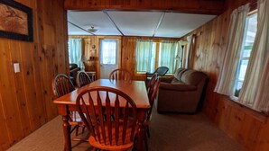 A wooden cabin interior with a dining table and chairs, a sofa, and a window with curtains. - Hawthorne Cottages Outpost (Raymond)