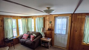 A cozy room with a brown sofa, a small table with a lamp, and a door with a window. - Hawthorne Cottages Outpost (Raymond)