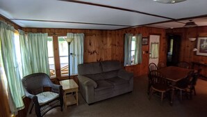 A cozy room with wooden walls, a grey sofa, a wicker chair, a small table, and a wooden dining table with chairs. - Hawthorne Cottages Outpost (Raymond)