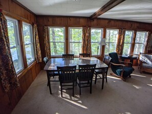 A wooden-paneled room with a round table and chairs, large windows with curtains, and a sofa. - Hawthorne Cottages Lakeside (Raymond)