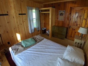 A wooden cabin bedroom with a bed, a dresser, a lamp, and a window with curtains. - Hawthorne Cottages Lakeside (Raymond)
