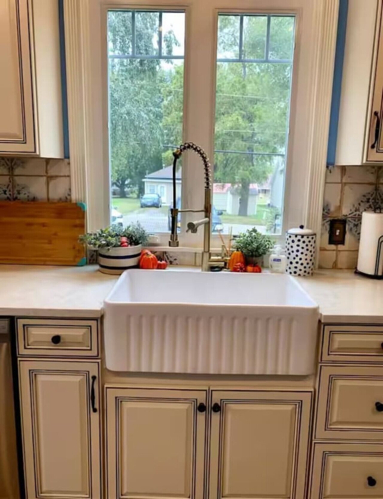 A kitchen with a white sink, beige cabinetry, and a window with a view of a house and trees.