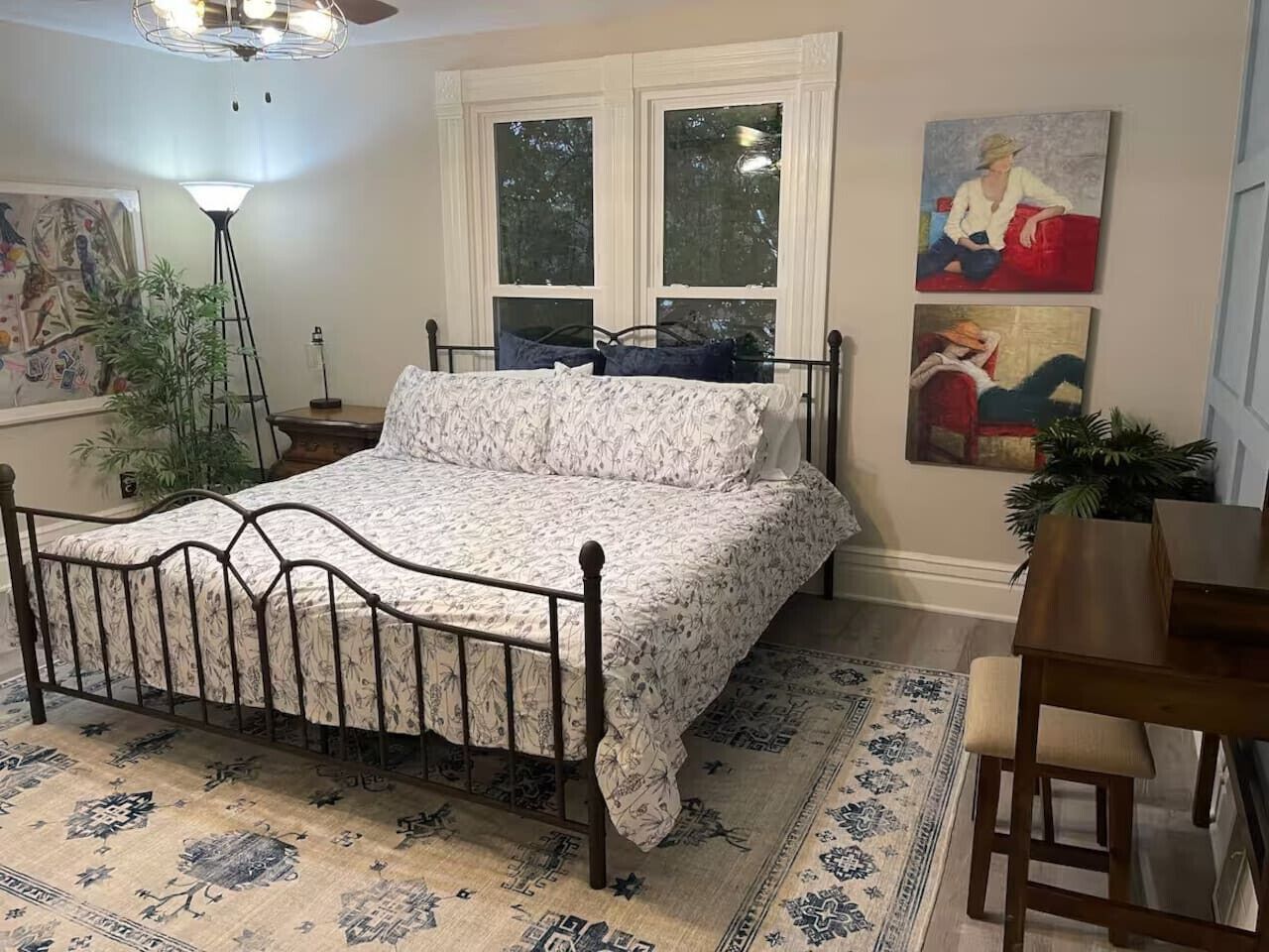 Un dormitorio con estructura de cama metálica, ropa de cama floral, una mesita de noche de madera, una lámpara de pie y una zona de comedor con mesa y sillas.