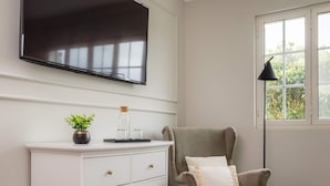Ein Zimmer mit weißem Schminktisch, einem grauen Sessel, einem flachbildfernseher an der Wand und einem Fenster mit Blick auf Grünflächen.