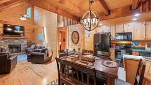 A rustic cabin interior with a wooden dining table, leather sofas, a fireplace, a kitchen with wooden cabinets, and a large TV mounted on the wall.