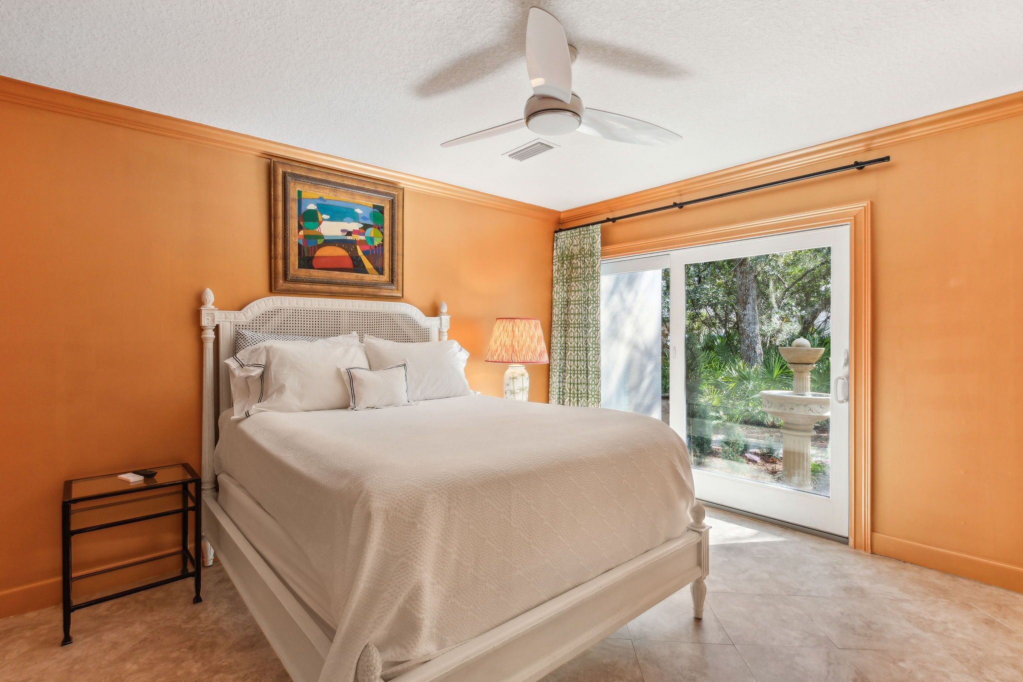 A bedroom with a bed, a ceiling fan, a nightstand, a lamp, and a window with a view of greenery.