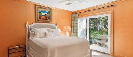 A bedroom with a bed, a ceiling fan, a nightstand, a lamp, and a window with a view of greenery.