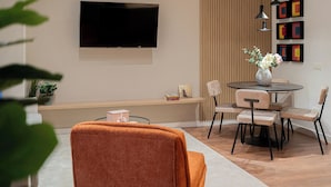 Una sala de estar moderna con un televisor de pantalla plana montado en la pared, una mesa de comedor redonda con sillas, un banco con libros y una planta en maceta.