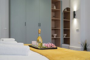 Una cama bien tendida con una manta amarilla, una botella de champán, una bandeja con flores rosadas y una toalla blanca.