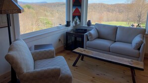 A cozy living room with a sofa, armchair, and a wooden coffee table. A window offers a view of a forested landscape. - Maison de LaMontagne: Unique hill top home with stunning views in Corinth (Corinth)