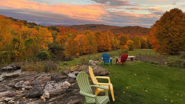 A scenic outdoor seating area with Adirondack chairs and a stone fire pit, overlooking a forested valley with autumn foliage. - Maison de LaMontagne: Unique hill top home with stunning views in Corinth (Corinth)