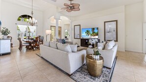 A spacious living room with a large sofa, a dining area with a chandelier, and a television displaying a tropical scene.