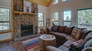 A cozy living room with a stone fireplace, a leather sofa, and a large window overlooking a deck. - Home with a Mountain View above the Angel Fire Golf Course (Angel Fire)