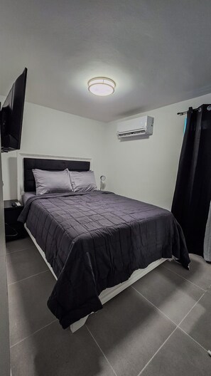 A bedroom with a bed, a flat-screen TV mounted on the wall, an air conditioning unit, and a curtain.