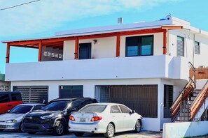 A two-story building with a garage, parked cars, and a staircase leading to the upper level. - Colonial Apartment w/ Parking & Patio, Near Beach (Carolina)