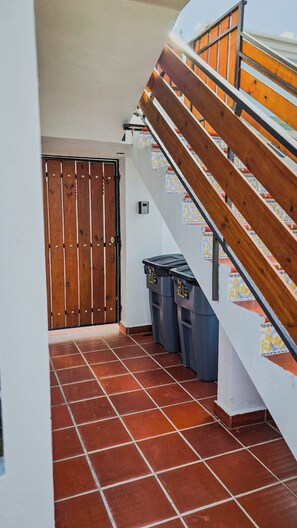 A narrow corridor with a wooden door, a staircase with patterned tiles, and two trash bins.