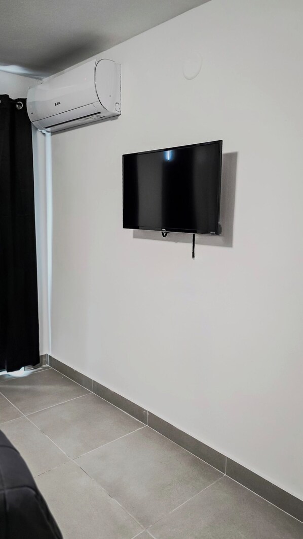 Una habitación con televisión de pantalla plana montada en la pared, aire acondicionado instalado en el techo y piso con baldosas.