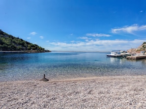 A clear view of a pebble beach with boats docked on the right, a calm sea, and a hilly landscape in the background.