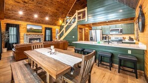 Interior rústico de una cabaña con comedor, chimenea, cocina con una isla grande y una escalera que conduce a un nivel superior.