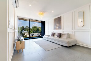 A modern living room with a white sofa, a chandelier, and a large window offering a view of the outdoors.