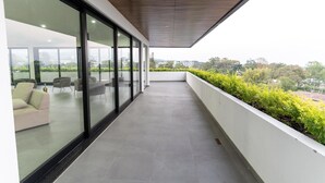 Ein moderner Balkon mit Glaswand, einer Sitzecke inklusive Sofa und Sesseln, und Blick auf Grünflächen sowie Gebäude in der Ferne.