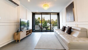 Ein modernes Wohnzimmer mit einem großen Flachbildfernseher, einem beigefarbenen Ledersofa, einem Holztische und einem großen Fenster mit Blick auf Palmen.