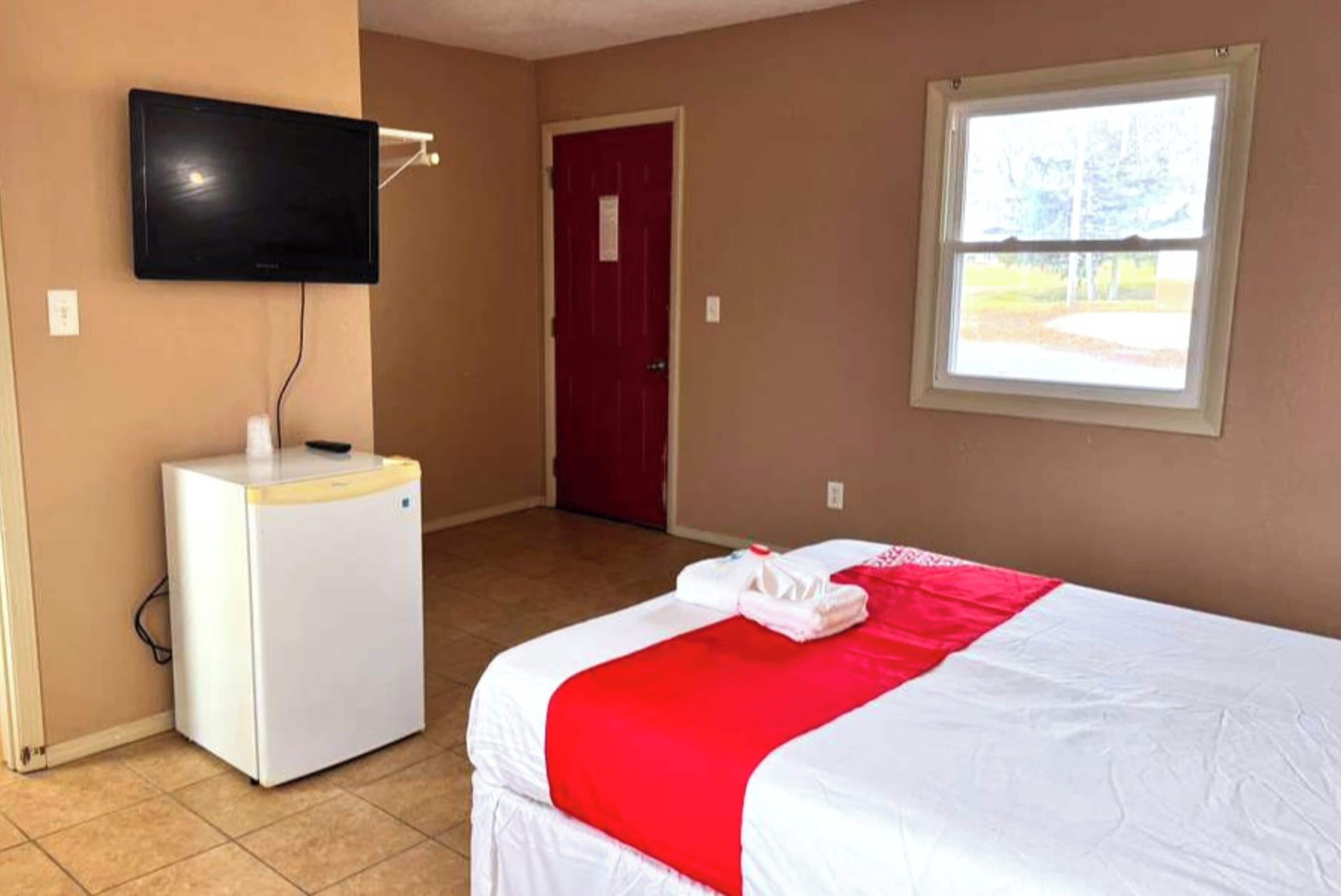 Habitación de hotel con cama, televisor montado en la pared, refrigerador pequeño, puerta roja y ventana con vista a árboles.