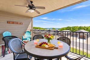 A round table with a fruit platter, drinks, and a vase of flowers on a balcony with chairs and a view of a residential area.