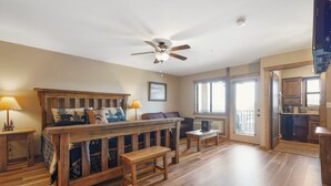 Een ruime woonkamer met een houten bed, een plafondventilator en uitzicht op de keuken.