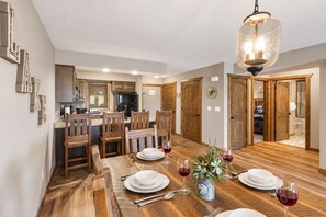 A dining area with a wooden table set for four, a chandelier, and a kitchen in the background.