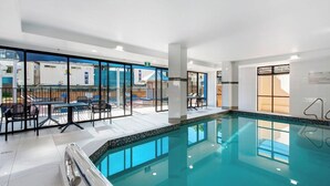 An indoor swimming pool with clear glass walls, a tiled floor, and a seating area with tables and chairs.