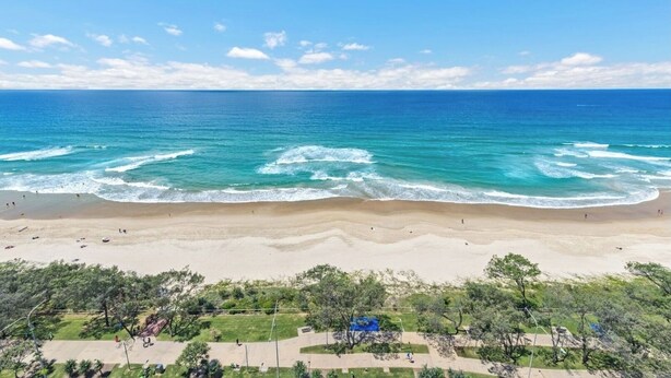 A beach with clear blue water, sandy shore, and greenery in the foreground.