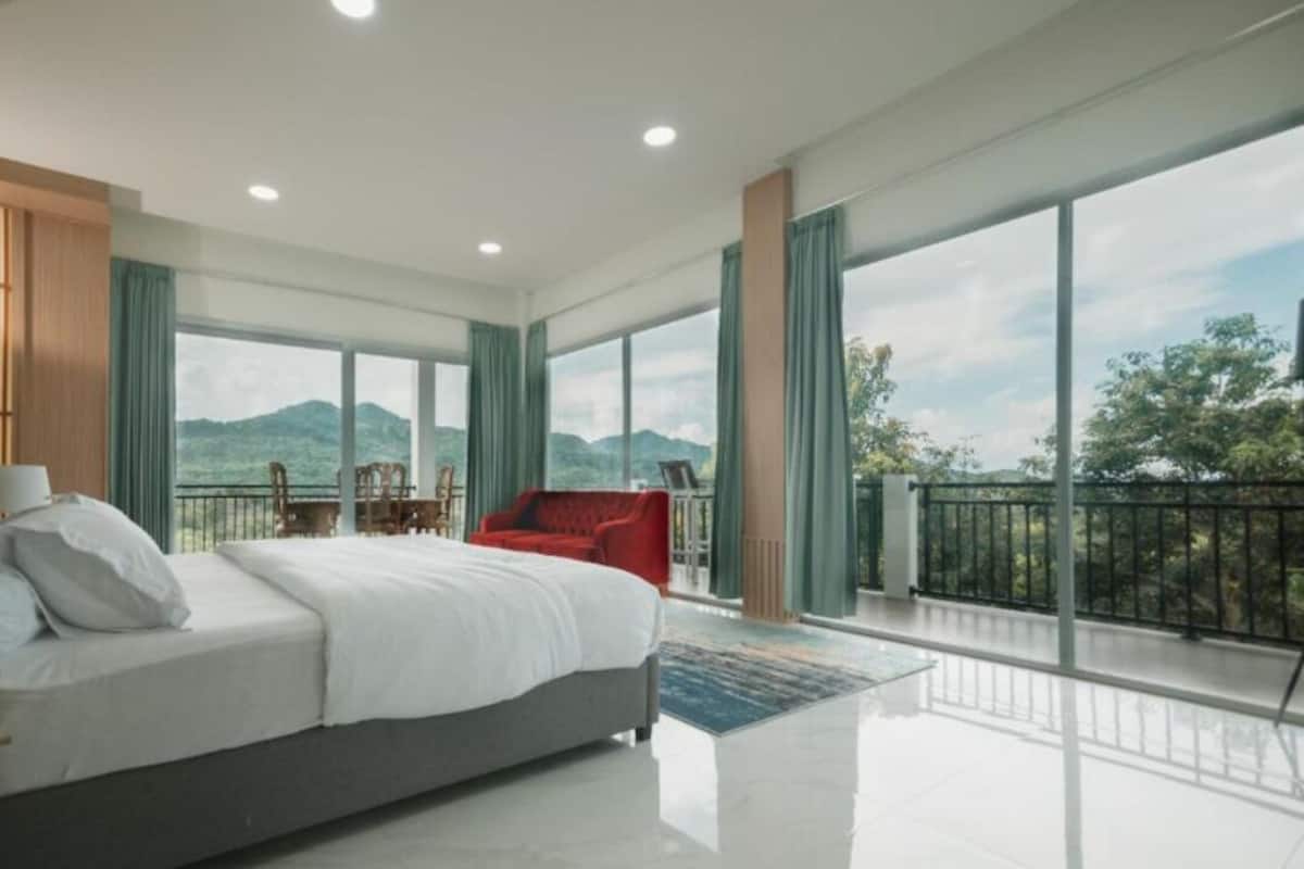 A spacious bedroom with a large bed, a red armchair, and a balcony with a mountain view.
