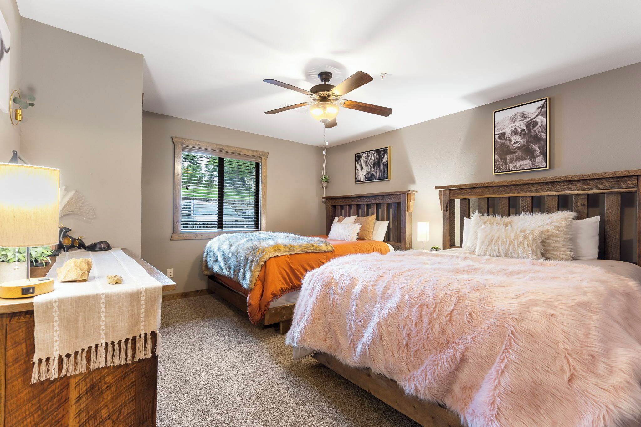 A bedroom with two beds, a ceiling fan, a window with blinds, a bedside table with a lamp, and a framed picture on the wall.