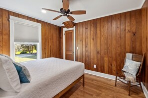 A bedroom with a bed, a ceiling fan, wooden walls, a window, and a chair. - Charming 2BR home in Saint Marys Historic Downtown: Dining, Shops, Ferry Steps Away (Saint Marys)
