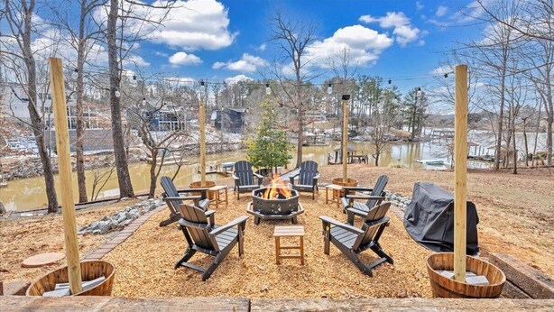 Un espace extérieur aménagé avec des chaises Adirondack disposées autour d’un foyer central, entouré de jardinières en bois et offrant une vue sur un lac et des bâtiments en arrière-plan.